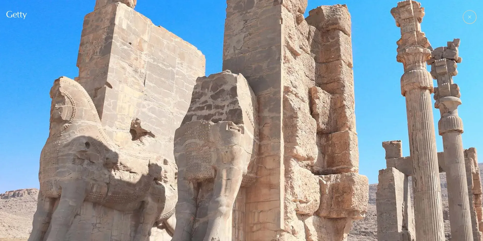 The present-day photograph that fills the screen after the petal frame is clicked, showing weathered and broken stone statues, door posts and several tall stone columns on the right under a blue sky