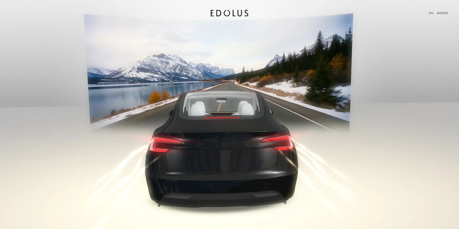 In an all-white space a black saloon stands in front of a large curved screen showing a road beside snowy mountains and a lake, with white streaks of light trailing from both sides of its rear